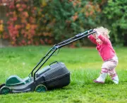 Cute curly baby girl in rain boots playing with a big green lawn mower in the garden